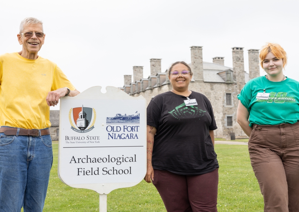 Three representatives standing by a sign reading Buffalo State Archaeological Field School sign at Old Fort Niagara